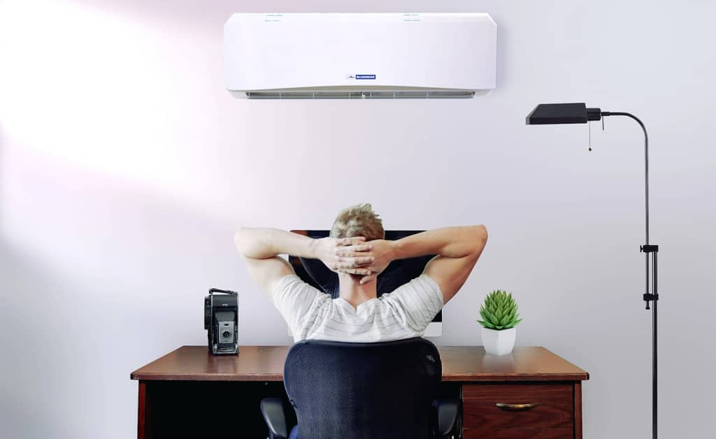 A man sitting at his desk with his back to the camera, hands behind his head, with a wall-mounted ductless mini-split AC above