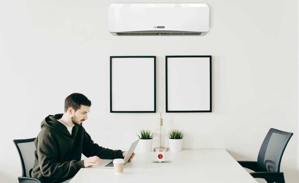 A man in a black jacket sitting at a desk with his laptop in front of him, a cup of coffee beside him, and a wall-mounted mini-split AC