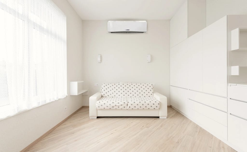 Wide room, big window left of white sofa, wall mounted mini-split AC behind. Built-in drawers and cabinets.