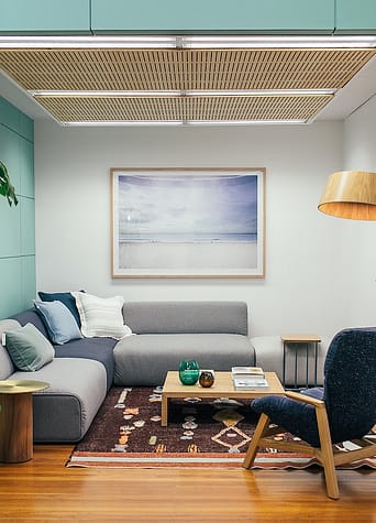 A living room with an L-shaped gray sofa, a wooden table in the center, a wooden blue chair, a portrait on the wall, and a ceiling cassette